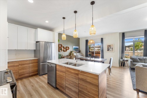 Modern kitchen featuring a large island with an undermount double sink, stainless steel appliances, two-tone cabinetry, wood-finish flooring, and recessed lighting - 6875 Evans Wynd Nw, Edmonton, AB - Indoor Photo Showing Kitchen With Double Sink