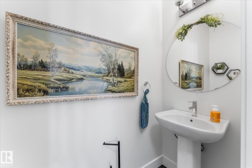 Pedestal sink with chrome faucet, circular wall mirror, and wall-mounted light fixture - 6875 Evans Wynd Nw, Edmonton, AB - Indoor Photo Showing Bathroom