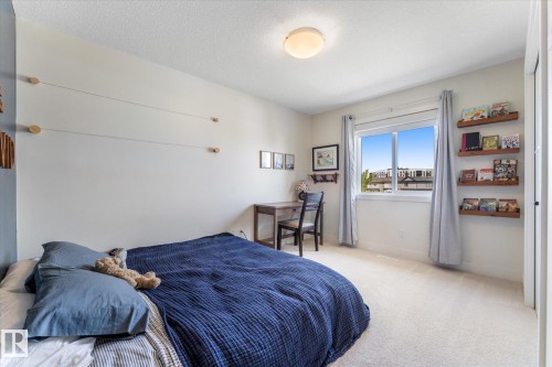 Carpeted bedroom featuring a large window, integrated wooden wall shelves, a built-in desk, and a flush-mount ceiling light fixture - 6875 Evans Wynd Nw, Edmonton, AB - Indoor Photo Showing Bedroom