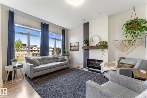 Bright living area featuring wood-finish flooring, a contemporary fireplace with a wood mantle, and large windows - 6875 Evans Wynd Nw, Edmonton, AB - Indoor Photo Showing Living Room With Fireplace
