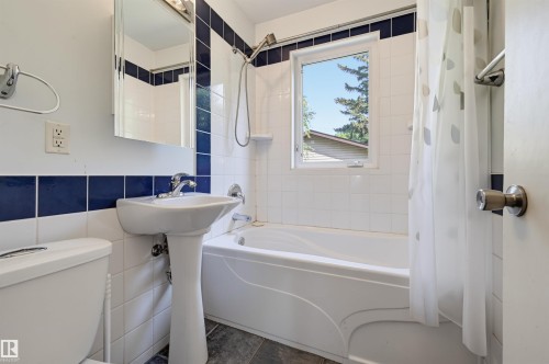 Bathroom featuring a pedestal sink, white tile walls with a blue accent band, a built-in bathtub with shower, and a framed window - 7754 80 Avenue Nw, Edmonton, AB - Indoor Photo Showing Bathroom