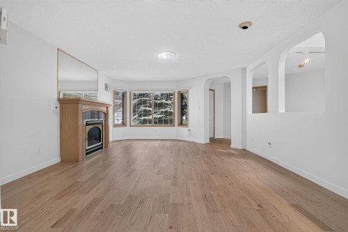 Spacious room featuring wood-finish flooring, a light wood mantel with a built-in fireplace, and a large bay window with outdoor scenery - 120 Ironwood Place Nw, Edmonton, AB - Indoor