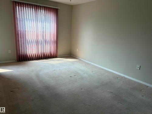 Room featuring neutral wall paint, beige carpet, white baseboards, and a large window with vertical blinds - #209 8912 156 St Nw, Edmonton, AB - Indoor Photo Showing Other Room
