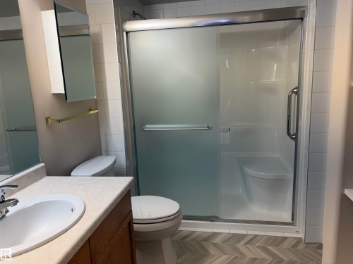 Bathroom featuring a sliding glass shower enclosure with an integrated seat, a vanity with a light-colored countertop and under-sink cabinetry, a toilet, a mirrored medicine cabinet, and herringbone pattern flooring - #209 8912 156 St Nw, Edmonton, AB - Indoor Photo Showing Bathroom