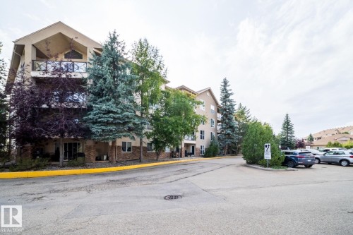 Multi-story residential building featuring balconies with glass railings, a stone facade on the lower levels, and mature evergreen trees - #209 8912 156 St Nw, Edmonton, AB - Outdoor