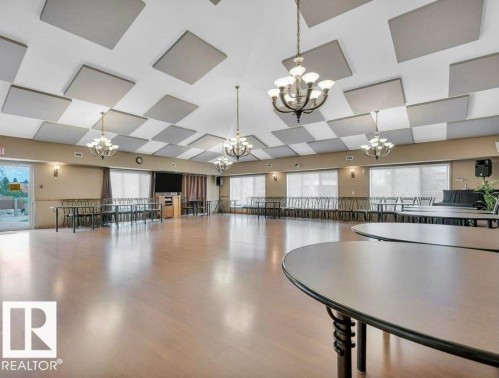 Spacious common area featuring light wood-finish flooring, multiple chandeliers, and large windows with roller blinds - #209 8912 156 St Nw, Edmonton, AB - Indoor Photo Showing Other Room