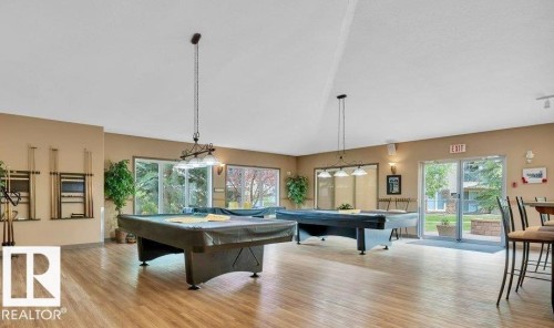 Expansive recreation room showcasing gleaming wood-finish flooring, dual billiard tables, abundant natural light from multiple windows, and pendant lighting fixtures overhead - #209 8912 156 St Nw, Edmonton, AB - Indoor Photo Showing Other Room