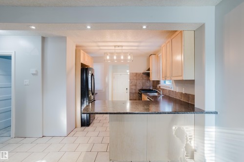 Kitchen featuring a built-in breakfast bar with a stone-finish countertop, light wood cabinetry, black appliances, recessed lighting, and a modern linear chandelier - 12431 81 Street Nw, Edmonton, AB - Indoor Photo Showing Kitchen