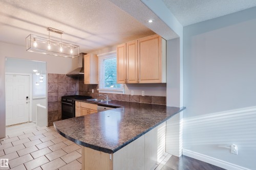Kitchen featuring a peninsula with a dark countertop, light wood-finish cabinetry, a stainless steel range hood, and a tiled backsplash - 12431 81 Street Nw, Edmonton, AB - Indoor Photo Showing Kitchen With Double Sink