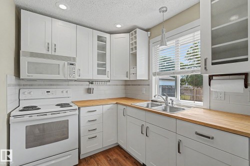 Kitchen featuring white cabinetry, wood-finish countertops, stainless steel double-basin sink, and a white oven with an over-the-range microwave - 8823 130A Avenue Nw, Edmonton, AB - Indoor Photo Showing Kitchen With Double Sink