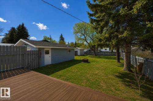 Spacious backyard with a composite deck, mature evergreen trees, a manicured lawn, and a detached building featuring blue siding and a white door - 4712 49 Street, Calmar, AB - Outdoor