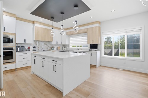 Gourmet kitchen featuring a large central island with a solid surface countertop, light wood-finish flooring, built-in stainless steel appliances, and a marble-finish backsplash - 304 33 Avenue Nw, Edmonton, AB - Indoor Photo Showing Kitchen With Upgraded Kitchen