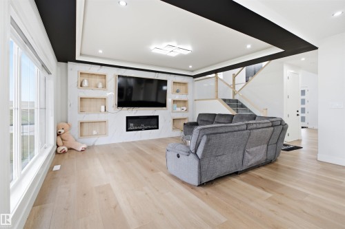 Open-concept living area featuring light wood-finish flooring, a contemporary fireplace with a stone-finish surround, recessed shelving, and a tray ceiling with integrated lighting - 304 33 Avenue Nw, Edmonton, AB - Indoor With Fireplace