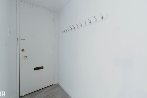 Entryway featuring a white door with brass hardware, a black mail slot, a wall-mounted coat rack with multiple hooks, white walls, and wood-finish flooring - #303 10883 Saskatchewan Dr Nw, Edmonton, AB - Indoor Photo Showing Other Room