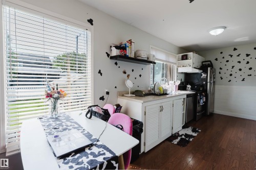 Kitchen area featuring wood-finish flooring, white cabinetry with louvered doors, a light-colored countertop, a stainless steel dishwasher, and a large window with blinds - 10955 72 Avenue Nw, Edmonton, AB - Indoor