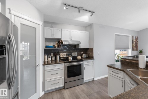 Modern kitchen featuring white cabinetry, stainless steel appliances, dark subway tile backsplash, wood-finish flooring, and a frosted glass pantry door - 4009 158 Avenue Nw, Edmonton, AB - Indoor Photo Showing Kitchen With Double Sink