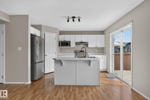Kitchen featuring an island with integrated sink and faucet, white cabinetry, light countertops, wood-finish flooring, and a sliding glass door to exterior - 8923 173 Avenue Nw, Edmonton, AB - Indoor Photo Showing Kitchen