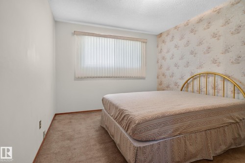 Carpeted room featuring a window with vertical blinds, light-colored walls, and one wall with floral wallpaper - 6012 145 Avenue Nw, Edmonton, AB - Indoor Photo Showing Bedroom