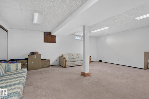 Expansive carpeted area featuring a drop ceiling with integrated fluorescent lighting, white painted walls, and a structural support column with a brick-finish base - 6012 145 Avenue Nw, Edmonton, AB - Indoor