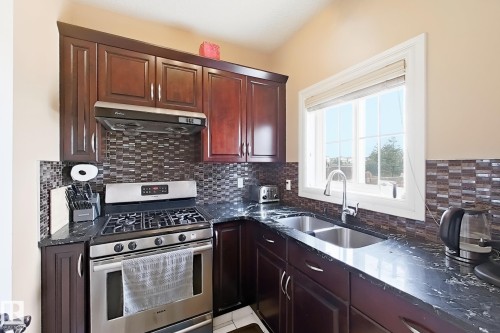 Kitchen featuring dark wood cabinetry, stainless steel oven with gas cooktop, mosaic tile backsplash, dark countertops, and a double basin sink beneath a window - 5931 12 Avenue Sw, Edmonton, AB - Indoor Photo Showing Kitchen With Double Sink