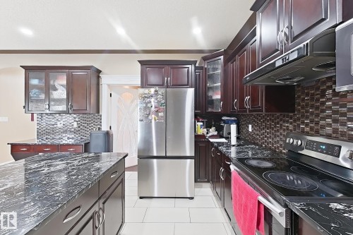 Kitchen featuring dark wood cabinetry, a large kitchen island with a dark stone countertop, a tile backsplash, and stainless steel appliances - 5931 12 Avenue Sw, Edmonton, AB - Indoor Photo Showing Kitchen With Upgraded Kitchen