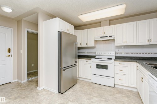 Kitchen featuring white cabinetry with gold-toned hardware, a stainless steel refrigerator, white electric range with overhead ventilation, dark countertops, and light-toned tile flooring - #413 237 Youville Dr E Nw, Edmonton, AB - Indoor Photo Showing Kitchen