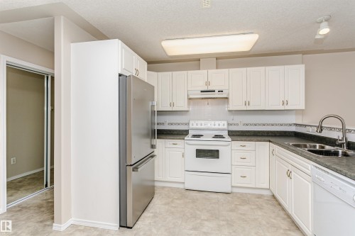 Kitchen featuring white cabinetry, dark countertops, a stainless steel refrigerator, white range, and a double basin sink - #413 237 Youville Dr E Nw, Edmonton, AB - Indoor Photo Showing Kitchen With Double Sink