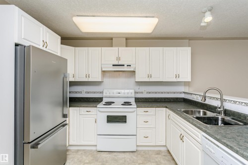 Kitchen featuring white cabinetry, dark countertops, and a full suite of appliances including a stainless steel refrigerator, white electric range, and integrated dishwasher - #413 237 Youville Dr E Nw, Edmonton, AB - Indoor Photo Showing Kitchen With Double Sink