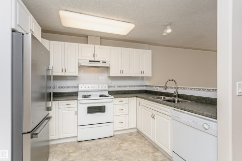 Kitchen featuring white cabinetry and appliances, a stainless steel refrigerator, a double basin sink with a gooseneck faucet, and patterned tile backsplash - #413 237 Youville Dr E Nw, Edmonton, AB - Indoor Photo Showing Kitchen With Double Sink