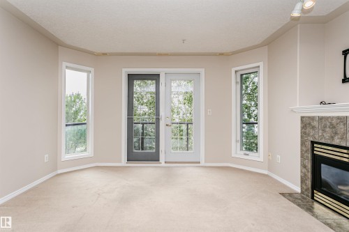 Carpeted living space featuring a fireplace with a tiled surround and white mantelpiece, recessed lighting, and a bay window configuration with a central glass panel door and two side windows - #413 237 Youville Dr E Nw, Edmonton, AB - Indoor With Fireplace
