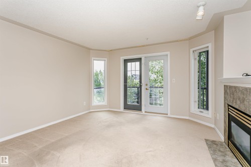Living area featuring light-toned carpeting, a fireplace with a tiled surround, a white mantel, and a glass-paned door opening to a balcony - #413 237 Youville Dr E Nw, Edmonton, AB - Indoor Photo Showing Other Room With Fireplace