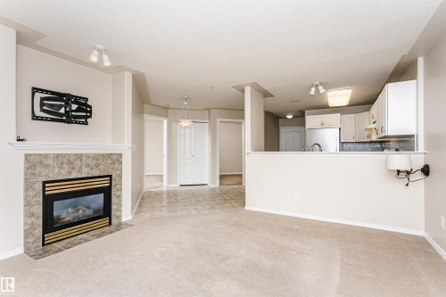 Living area featuring a tiled fireplace with mantel, beige carpeting, an open kitchen pass-through, and neutral wall tones - #413 237 Youville Dr E Nw, Edmonton, AB - Indoor Photo Showing Living Room With Fireplace