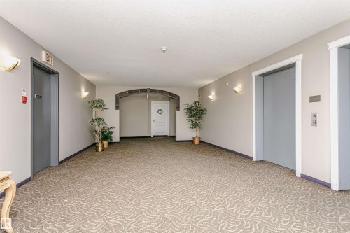 Hallway featuring patterned carpet flooring, light-colored walls, multiple wall sconces providing ambient lighting, and two elevators with gray doors - #413 237 Youville Dr E Nw, Edmonton, AB - Indoor Photo Showing Other Room