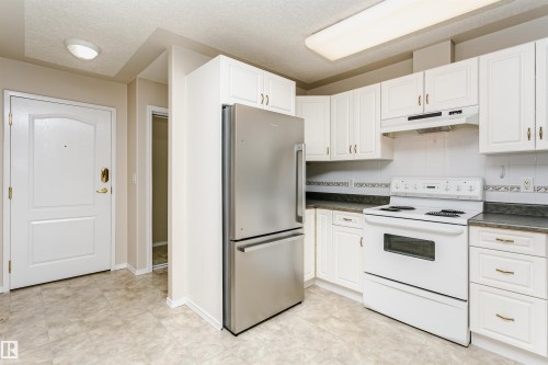 Kitchen featuring white cabinetry, a stainless steel refrigerator, white electric range with overhead ventilation, dark countertops, and a patterned tile backsplash - #413 237 Youville Dr E Nw, Edmonton, AB - Indoor Photo Showing Kitchen