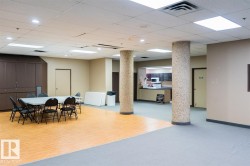 Spacious common area featuring mixed flooring with wood-finish sections and carpeting, two textured support columns, and a dedicated kitchenette with dark cabinetry and white countertops - 