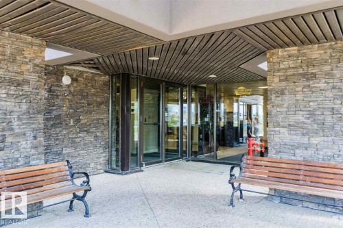 Stone facade entry featuring two wood benches with cast iron frames, glass double doors, a wood-finish slatted ceiling, and an exterior wall light fixture - #509 10149 Saskatchewan Dr Nw, Edmonton, AB - Outdoor With Deck Patio Veranda