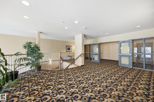 Spacious interior corridor with patterned carpet flooring, recessed ceiling lighting, a wood and metal railing system, and multiple glass-paneled doors - #443 10121 80 Av Nw, Edmonton, AB - Indoor Photo Showing Other Room