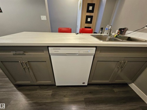 Kitchen island featuring a double basin stainless steel sink, a white Amana dishwasher, and gray shaker-style cabinetry with silver hardware, all complemented by wood-finish flooring - 13309 104 Street Nw, Edmonton, AB - Indoor Photo Showing Other Room