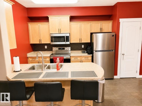 Kitchen featuring wood-finish cabinetry, stainless steel appliances, a double basin sink, and a breakfast bar - #202 16715 100 Av Nw, Edmonton, AB - Indoor Photo Showing Kitchen With Double Sink