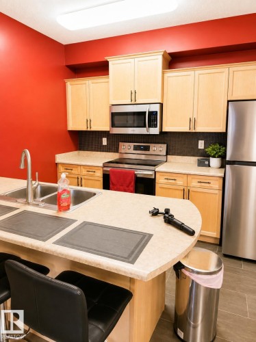 Kitchen featuring light wood-finish cabinetry, stainless steel appliances, a dark mosaic tile backsplash, and an island with a double basin sink - #202 16715 100 Av Nw, Edmonton, AB - Indoor Photo Showing Kitchen With Double Sink