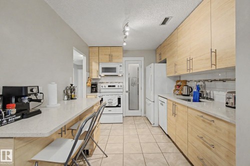 Light wood-finish cabinetry with brushed nickel hardware, white quartz countertops, full-size white appliances, tile flooring, and a glass-paneled pantry door - #39 3111 142 Av Nw, Edmonton, AB - Indoor Photo Showing Kitchen
