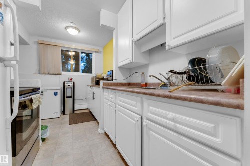 Galley kitchen featuring white cabinetry, light-toned flooring, and a window with blinds - #104 11445 41 Av Nw, Edmonton, AB - Indoor Photo Showing Kitchen