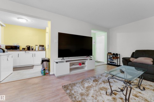 Open-concept living area featuring wood-finish flooring, a white media console, and a distinct opening to a kitchen area with white cabinetry and yellow accent walls - #104 11445 41 Av Nw, Edmonton, AB - Indoor Photo Showing Living Room