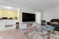 Open-concept living area featuring wood-finish flooring, a white media console, and a distinct opening to a kitchen area with white cabinetry and yellow accent walls - 