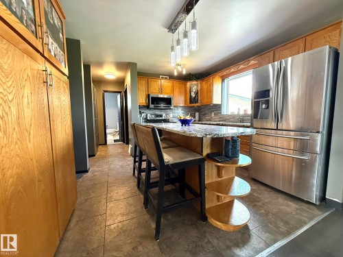 Kitchen featuring wood cabinetry, stainless steel appliances, a multi-tone backsplash, and a granite-finish island with integrated shelving - 4508 57 Avenue, Lamont, AB - Indoor