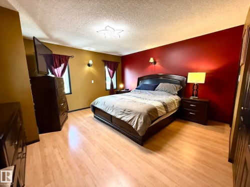 Bedroom featuring light wood-finish flooring, accent wall, two windows with dark drapery, and a decorative ceiling light fixture - 4508 57 Avenue, Lamont, AB - Indoor Photo Showing Bedroom