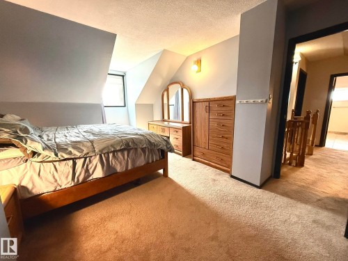 Carpeted room featuring a sloped ceiling, a window with dark trim, a wall-mounted light fixture, and a doorway opening to a hallway with wood railing - 4508 57 Avenue, Lamont, AB - Indoor Photo Showing Bedroom