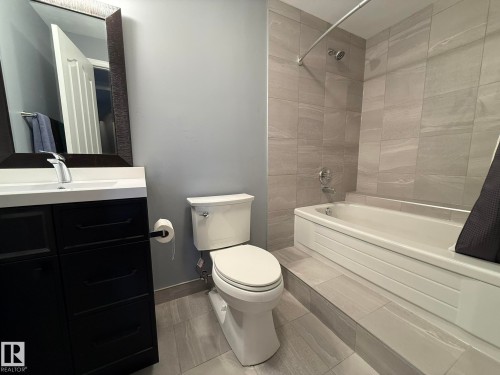 Bathroom featuring a tub/shower combination with light gray tile surround, a white vanity with integrated sink, black cabinetry, and a framed mirror - 4508 57 Avenue, Lamont, AB - Indoor Photo Showing Bathroom