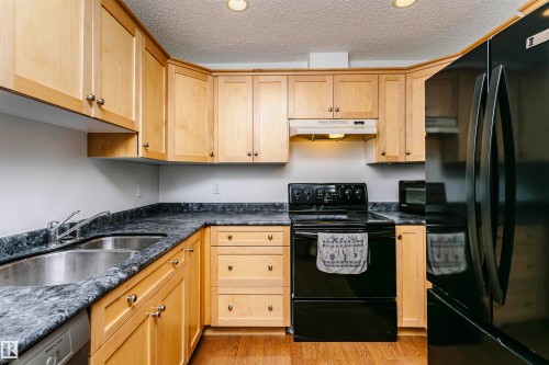 Kitchen featuring light wood cabinetry, black appliances, a double basin stainless steel sink, dark patterned countertops, and wood-finish flooring - #106 12110 106 Av Nw, Edmonton, AB - Indoor Photo Showing Kitchen With Double Sink