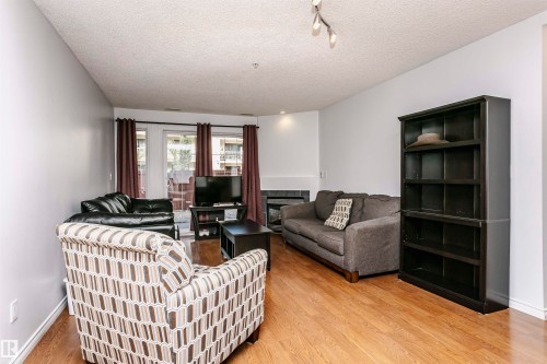 Living room featuring wood-finish flooring, light gray walls, and a corner fireplace with a dark tile surround and white mantel - #106 12110 106 Av Nw, Edmonton, AB - Indoor Photo Showing Living Room